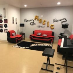 A music room at an ICF/DD home in Reseda. It features red sofas and chairs, a digital piano with a bench, music stands, and wall decorations with musical notes and instruments. There is also a piano-key rug and an LP art wall.